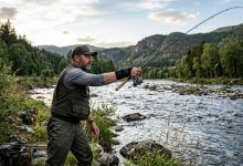 An angler wearing a black wrist brace while casting a fishing rod from a riverbank